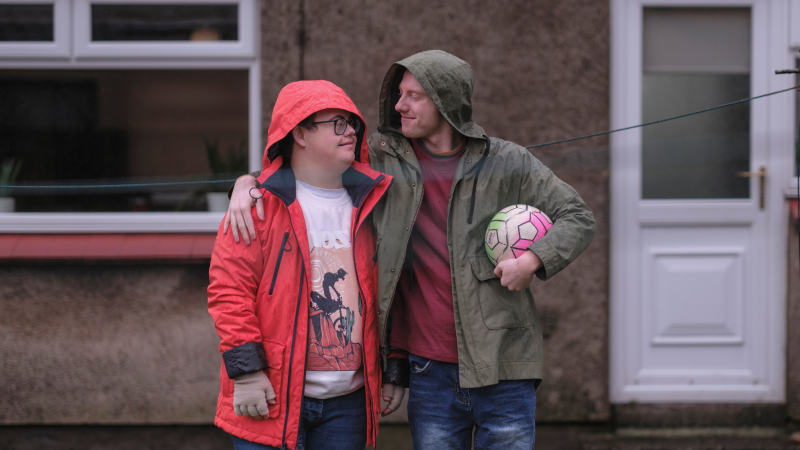 Two people standing outside a house sharing a hug with one holding a football under his arm