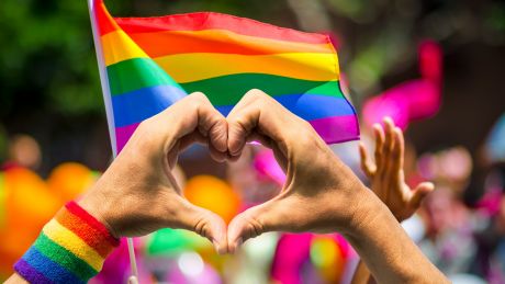 Person making heart symbol with their hands with pride flag in the background